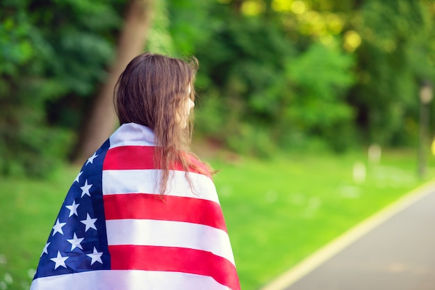 Back view of  young woman posing with usa national flag standing outdoors at green forest