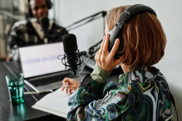 Photo back view of young woman recording podcast in studio and wearing headphones