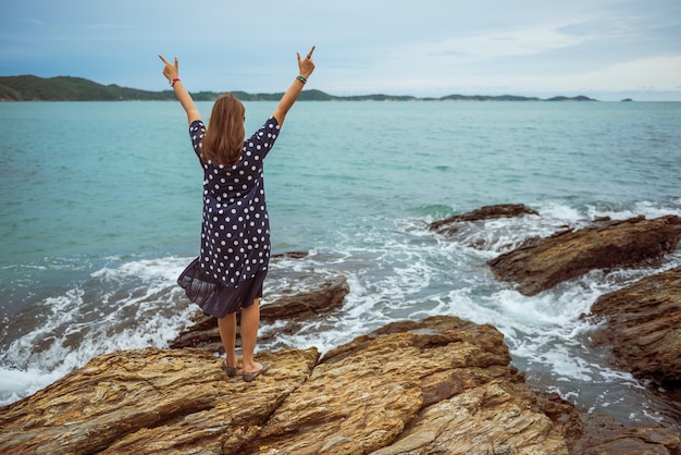 Back view of young woman standing on the rock raised hands up at the beach.