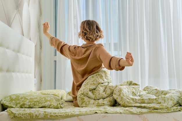 Back view of young woman stretching arms sitting on the bed at home bedroom looking to the window