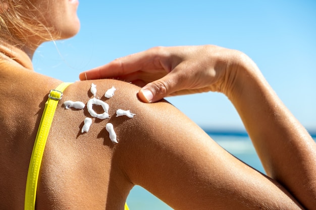 Вид сзади молодой женщины, загорающей на пляже с солнцезащитным кремом в форме солнца на ее плече.