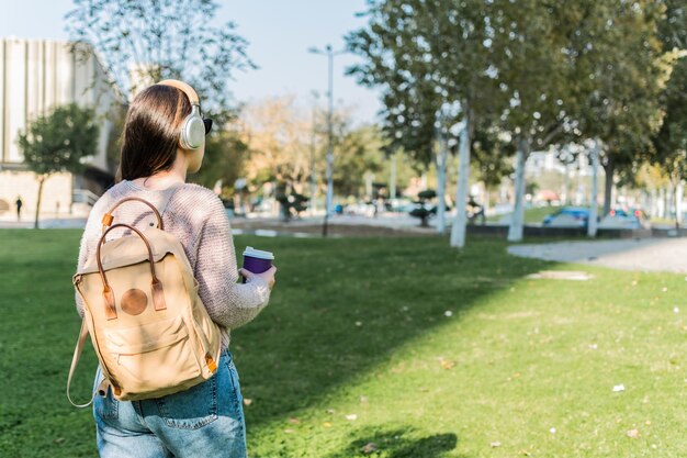 晴れた日に公園を歩くバックパックコーヒーとヘッドフォンを持つ若い女性の背面図