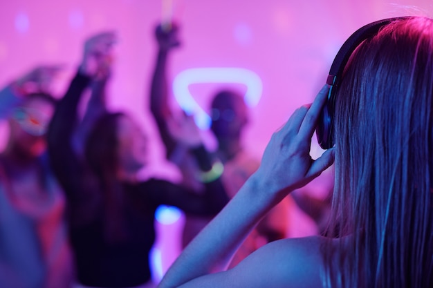 Back view of young woman with long blond hair touching headphones while standing in front of crowd of excited friends dancing at party