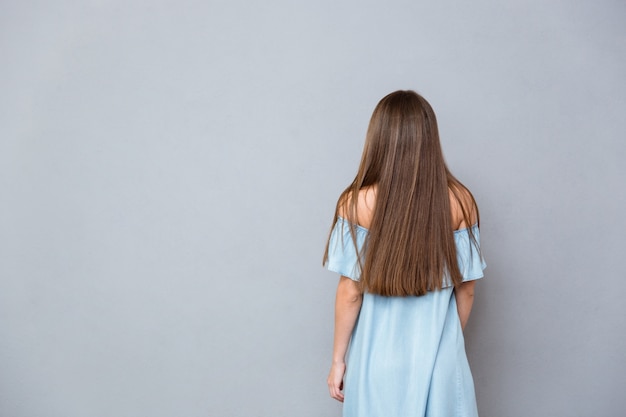 Photo back view of young woman with long hair in blue dress standing on gray wall