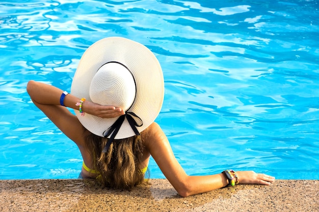 晴れた日に青い水と暖かい夏のプールでリラックスした黄色の麦わら帽子をかぶった長い髪の若い女性の背面図。