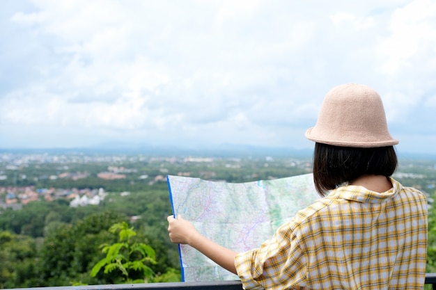 Back of young asian girl traveler holding map while standing in outdoors nature background