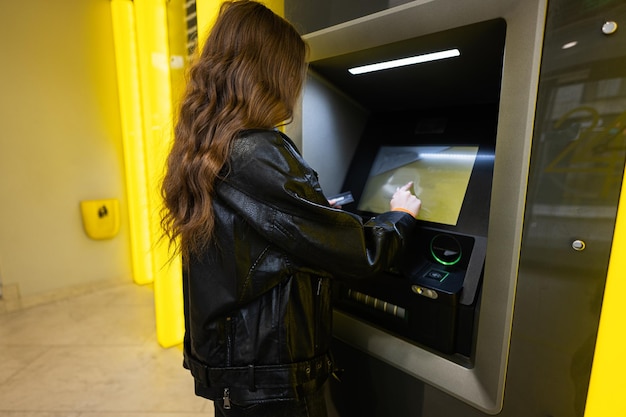 Back of young brunette girl withdrawing money from credit card at yellow ATM