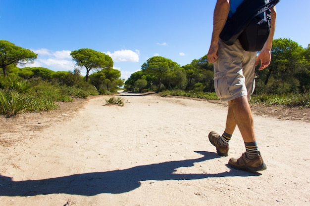 Back of young man in shorts and t shirt with camera bag walking on sand path in national park Cadiz