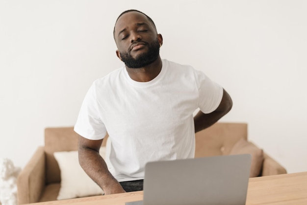 Backache African american man suffering from low back pain from strenuous work on laptop while sitting at workplace in modern home office Lumbar pain Health problem concept