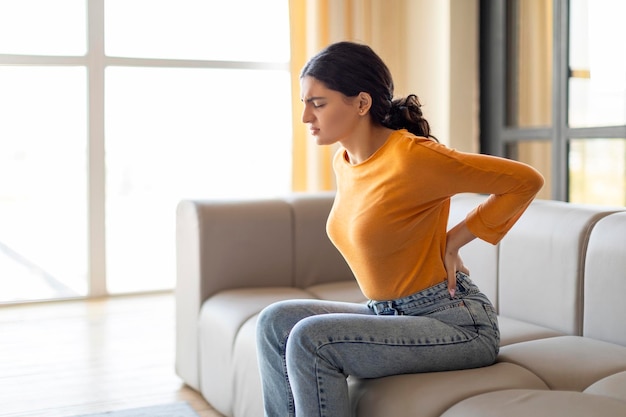 Foto concept di mal di schiena giovane donna indiana che massaggia il mal di schiena mentre è seduta sul divano