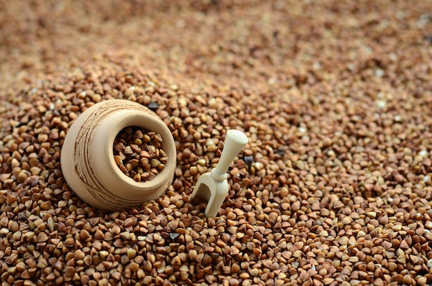 Background image of a large pile of buckwheat, in the middle of which lies a small jug and a wooden spatula for cereals