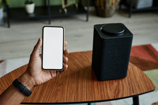 Background image of male hand holding phone with blank screen next to home AI smart speaker