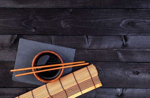 Background for sushi. Bamboo mat and soy sauce on black wooden table. Top view with copy space