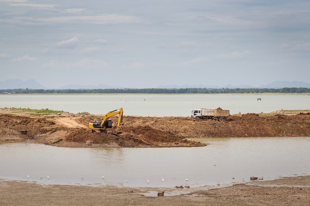 Photo backhoe (excavators) and trucks in the area of construction with river and sky as a backdrop.