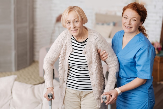 Поддержка пожилых людей. Оптимистичная активная красивая женщина, использующая костыли, в то время как прекрасная медсестра держит ее за руку и поддерживает