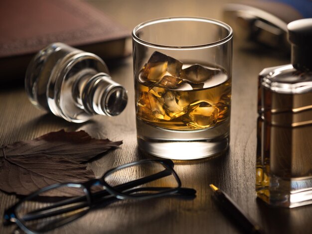 Backlit glass of whisky on the rocks on a wooden table with notebook pen and reading glasses