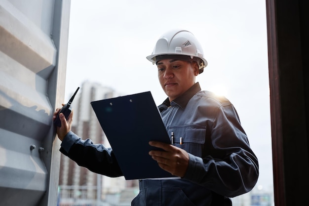 Backlit portrait of female worker wearing hardhat standing in\
container door at shipping docks