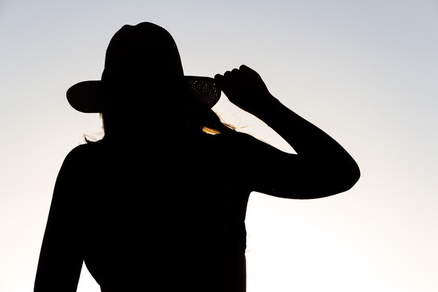 Backlit silhouette of a woman holding a hat with one hand.