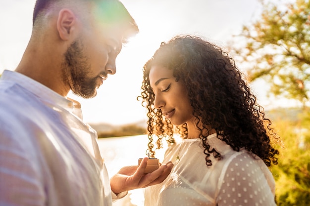 Foto scena romantica retroilluminata suggestiva di una giovane bella coppia multirazziale innamorata. bel ragazzo che fa la proposta di matrimonio alla sua ragazza ispanica nera che mostra l'anello di fidanzamento vicino a lei