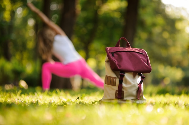 Рюкзак на траве в зеленом парке на летний день против женщины делают йоги