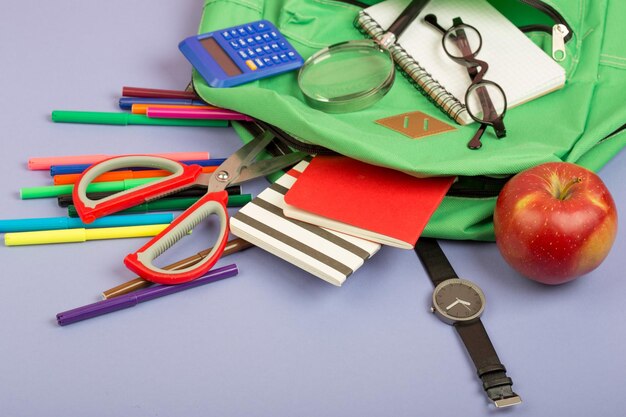 Backpack and school supplies magnifying glass notepad felttip pens eyeglasses scissors calculator watch on blue paper background