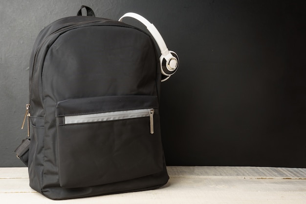 Backpack with headphones on a black wall, on a white wooden table copy space