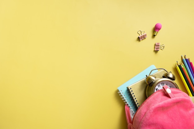 Рюкзак с блокнотами и будильником с плюшевым мишкой и красочными карандашами на цветном фоне с копией пространства Вернуться к школьной концепции