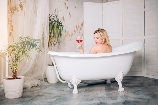 Backside portrait of naked female model sitting in warm bath with glasses of red wine enjoying spa procedure in sunny day, relaxing in modern luxury bathroom with green plants in hotel or at home.