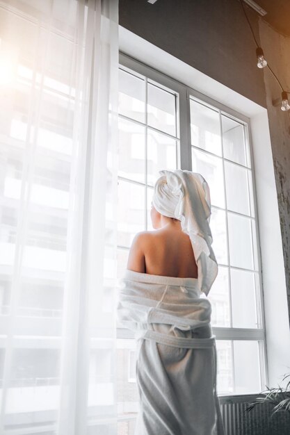 Backside portrait of slim caucasian woman with white towel on head standing against the big window in bathroom after having bath, taking off her bathrobe, showing bare shoulders.