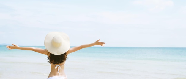 Backside young woman skin tan in sunhat and bikini standing with her arms raised to her head enjoying on the beach vacation travel and enjoy life at sea looking view of beach ocean on hot summer day