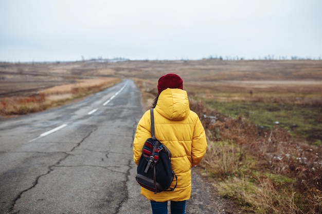 Backview van een vrouwelijke toerist met een rugzak die gele jas en rode hoed draagt, loopt op de weg.