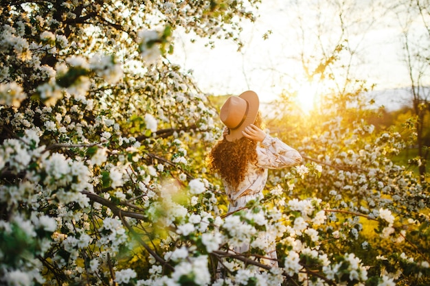 Backview of a young woman among the white blossom of the apple trees at the spring park. Female wearing white dress and beige hat feeling fresh and enjoys good weather on the sunset. Season concept.