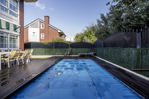 Backyard of a singlefamily house with a swimming pool covered by a blue protective tarp acacia wood floors a metal perimeter fence dining table with matching chairs