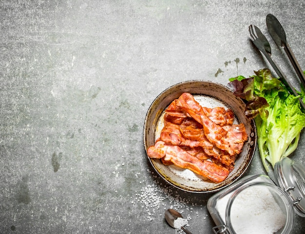 Bacon in a frying pan with salt and herbs. On a stone background.