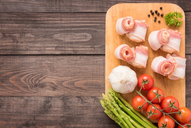Bacon rolls with tomato, garlic, asparagus on wooden background