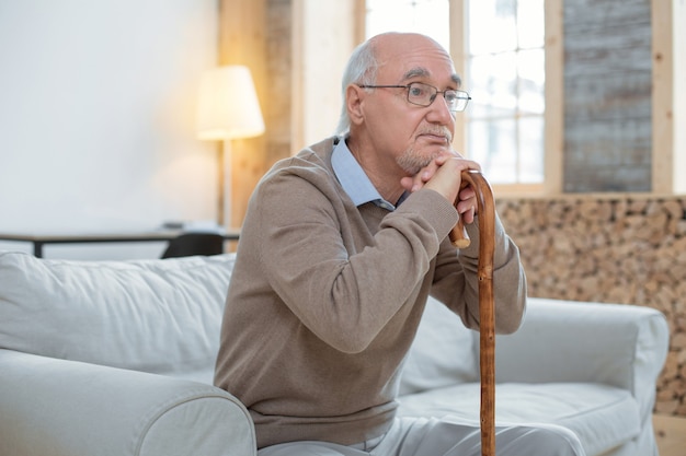 Bad day. Attractive thoughtful senior man leaning on cane while wearing glasses and sitting on couch