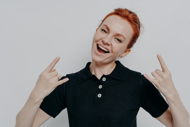 Bad girl. Cheerful carefree ginger female in casual clothes showing heavy metal rock sign while posing isolated on grey background in studio, smiling at camera rock and doing roll gesture
