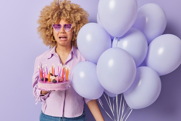 Bad holiday concept Upset crying woman with curly hair holds festive cake with candles bunch of inflated balloons wears sunglasses and shirt isolated over purple background celebrates birthday alone