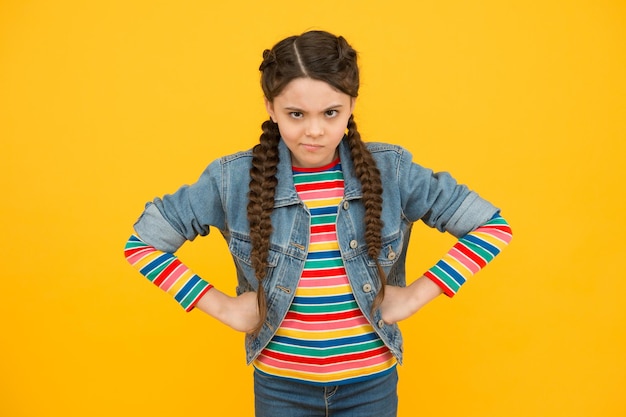 Photo in bad humor. moody little child yellow background. little baby wear braided hairstyle. little girl in casual style. little kid with hands on hips. trendy fashion. stylish trends. hair salon.