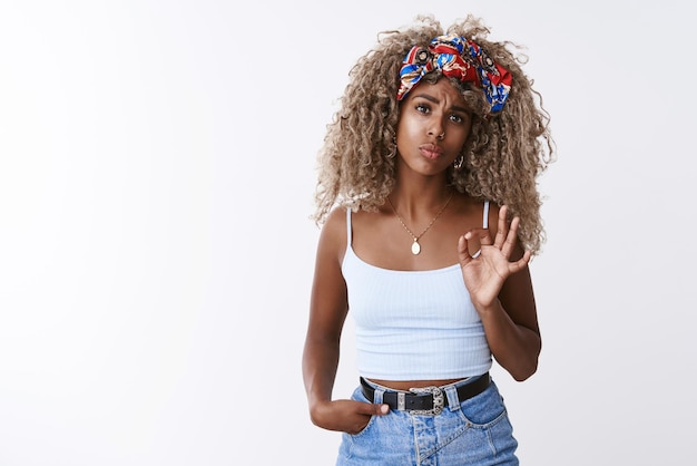 Not bad for newbie Impressed sassy and stylish hipster modern africanamerican woman with blond curly hair folding lips frowning astonished show okay fine good gesture white background