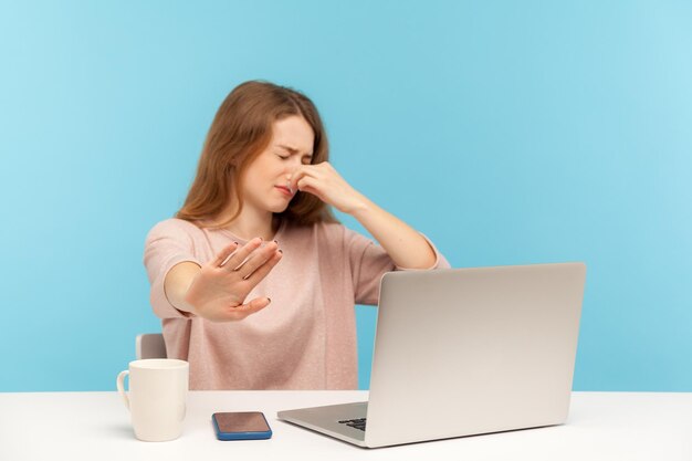 Bad smell. Confused young woman employee sitting at workplace with laptop and holding breath, pinching her nose to avoid awful stink, gesturing stop. indoor studio shot isolated on blue background