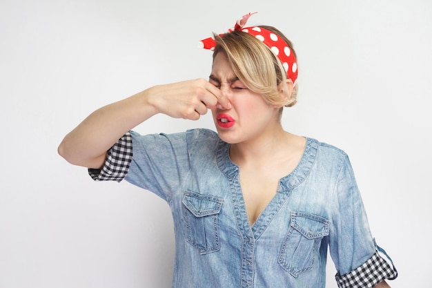 Bad smell. Portrait of dissatisfied beautiful young woman in casual blue denim shirt with makeup and red headband standing pinching her nose. indoor studio shot, isolated on white background.