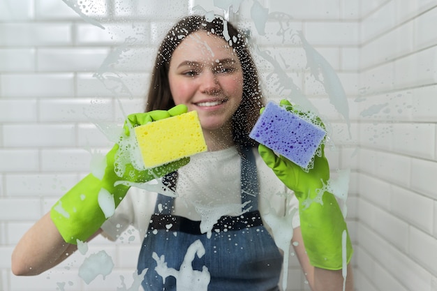 Badkamer schoonmaken, tienermeisje wassen doucheglas met schuim en sponzen, close-up van vrouwtjes hand in groene handschoenen en gekleurde sponzen