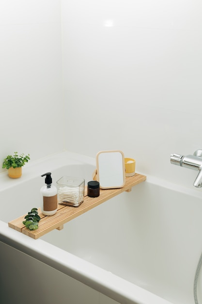 Badkuip houten dienblad met cosmetische schoonheidsflessen en accessoires in een moderne Scandinavische badkamer met witte tegels als achtergrond. Home decor met kaarsen en groene planten.