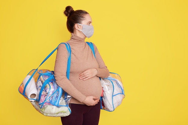 Bags of baby clothes, stuff, pregnant woman being ready for going to maternity hospital, lady wearing casual clothing and medical mask, giving birth during corona virus.