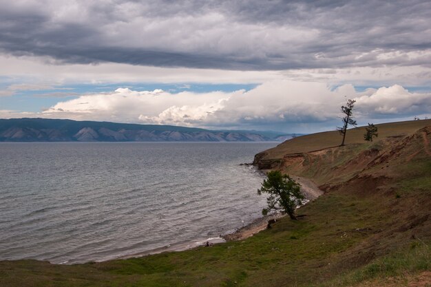 背景に山々と美しい雲があるバイカル湖畔。湖岸の木々。波のある湖。