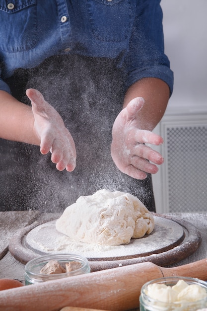 Bakconcept, strooi bloem op gistdeeg. Meel, melk en eieren op houten snijplank, gebakje ingrediënten. Bijgesneden afbeelding van onherkenbare vrouw kneden. Vrouwelijke bakkersclose-up, verticaal