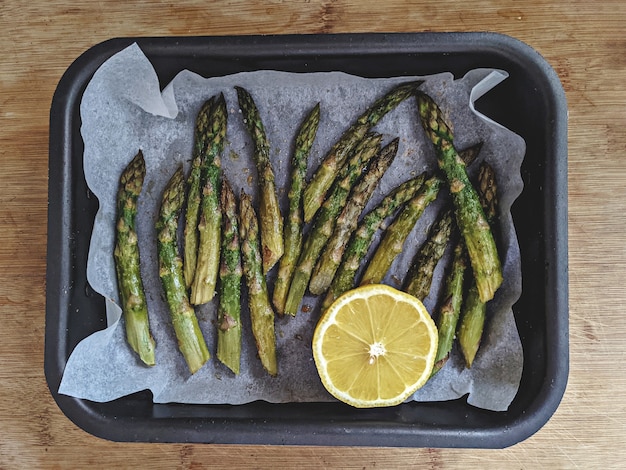 Baked asparagus in baking dish cooking in oven and half of lemon on wooden surface table