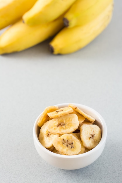 Фото Запеченные банановые чипсы в белой миске и связка бананов на столе. быстрое питание. скопируйте пространство. вертикальный вид