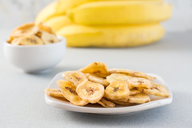 Chips di banana al forno in una ciotola e piattino bianchi e un mazzo di banane sul tavolo. fast food.